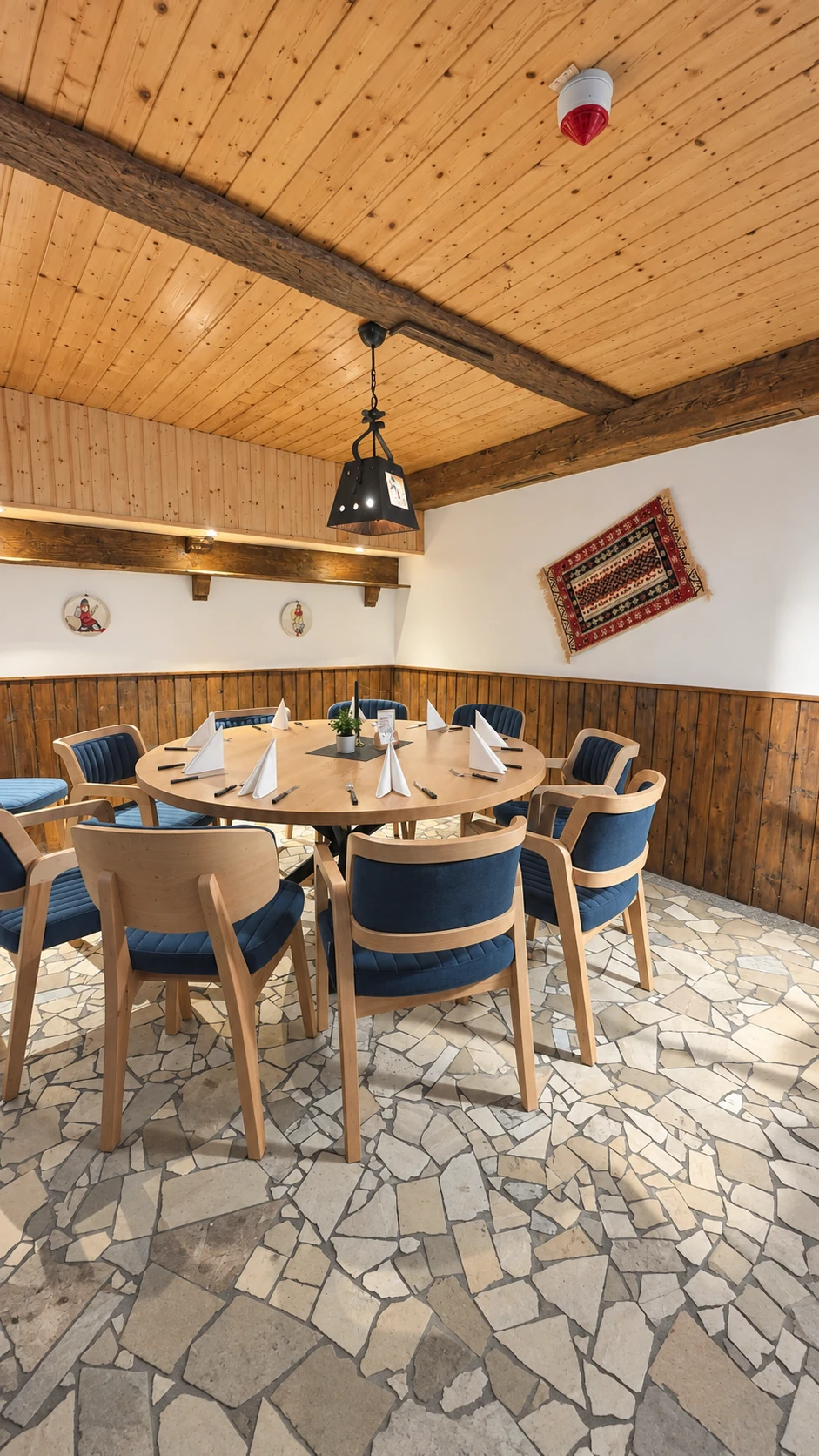 Holzvertäfelte Stube im Hotel Hasa mit rundem, eingedecktem Tisch und albanischem Kelim an der Wand.